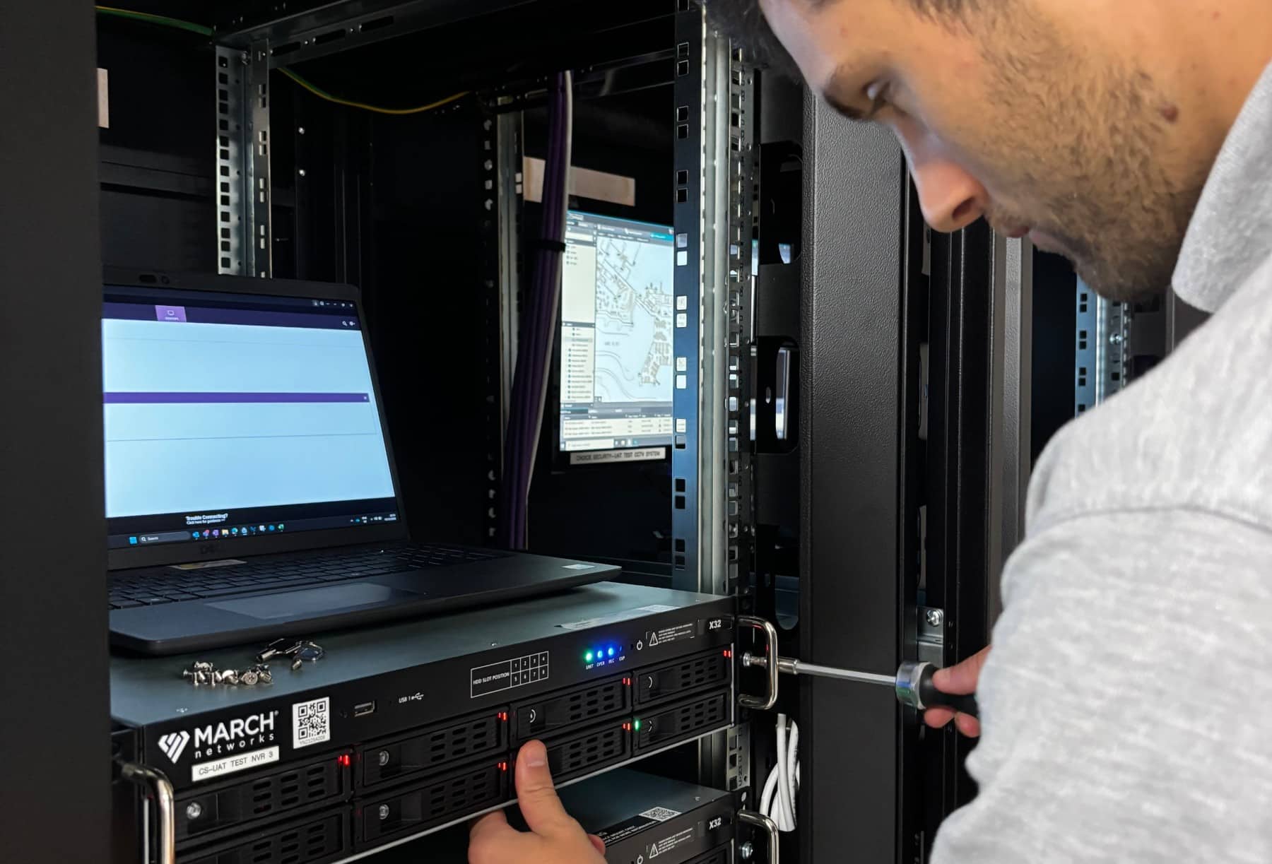 Man working with server equipment in a data center, using a screwdriver and a laptop.