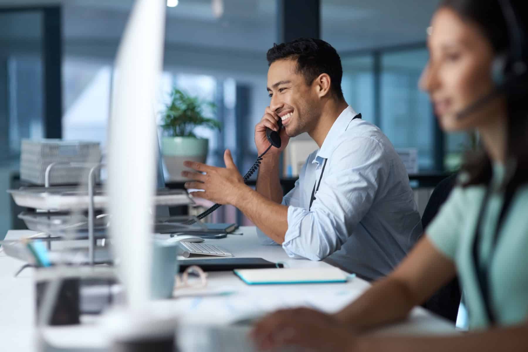 Smiling man talking on the phone at an office desk with a coworker nearby.