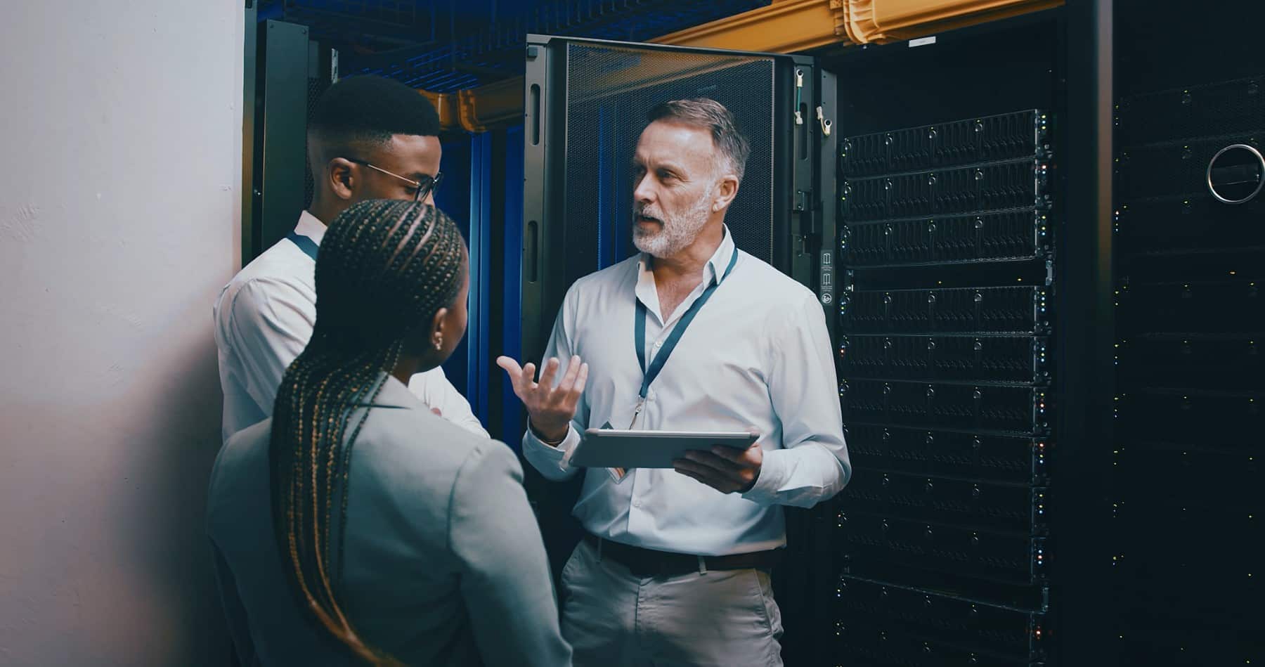 Three people have a discussion in a server room, one holding a tablet and explaining something.