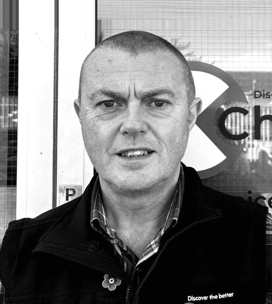 MarkBettles A man with short hair stands outside, wearing a jacket and shirt, looking at the camera.