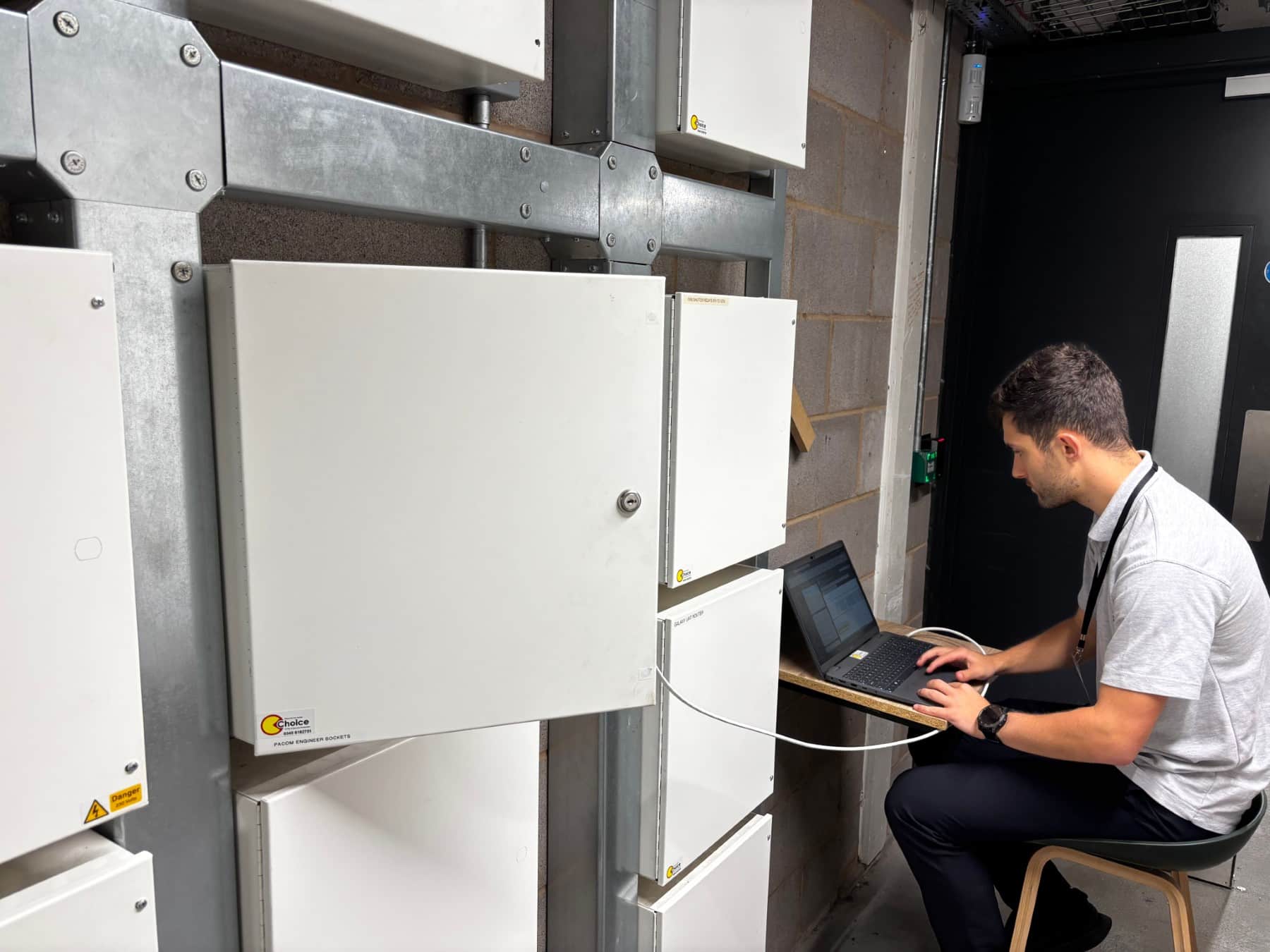 A technician sits at a laptop connected to control panels on a wall in a utility room.
