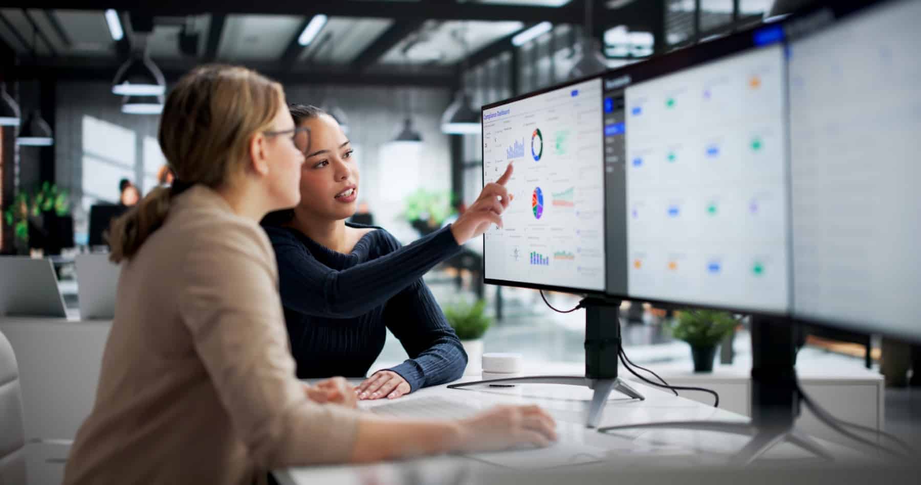 Tailored SLAs Bespoke Reporting Two women looking at data and charts on computer monitors in a modern office setting.