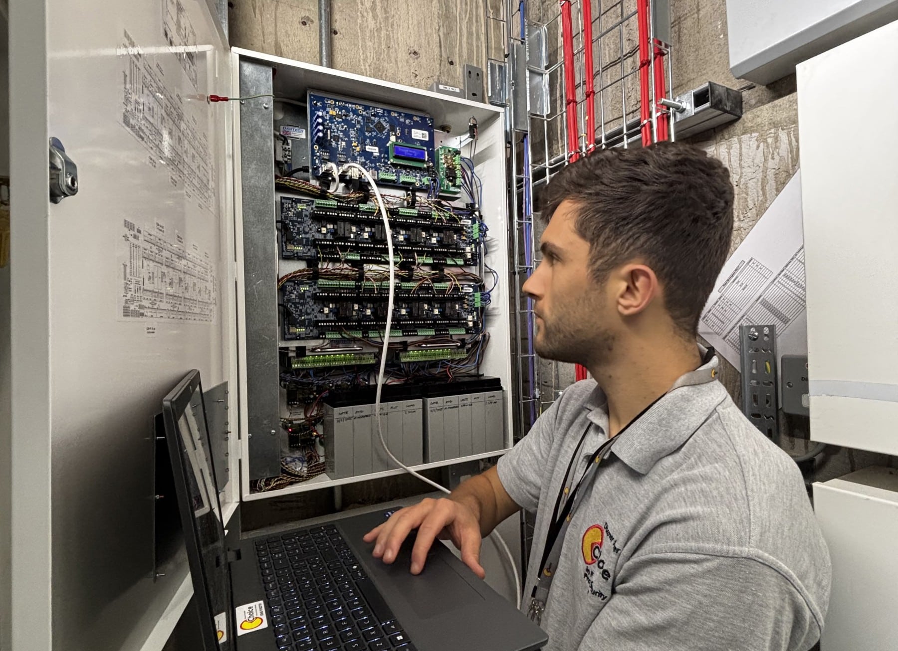 Man working on a laptop next to an open electrical control panel in a building.